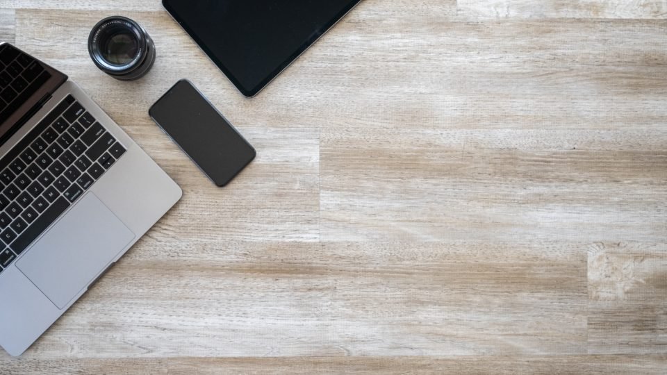 Notebook, tablet, celular e uma lente de câmera sobre mesa de madeira clara, vistos de cima